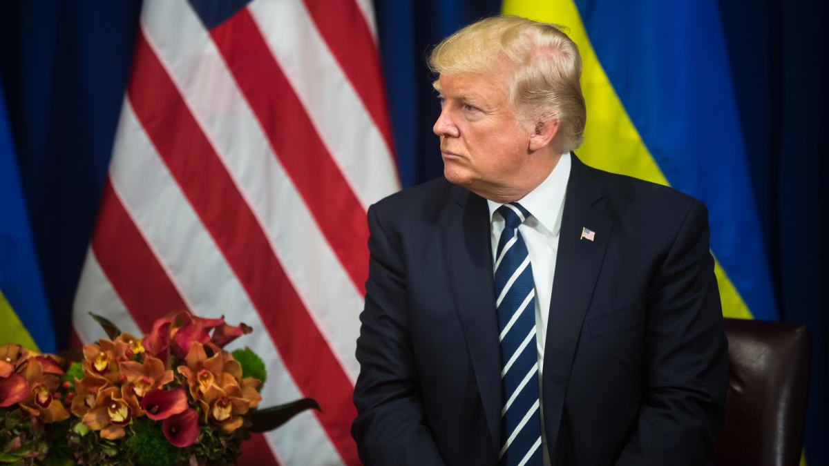 A man in a dark suit and striped tie sits at a desk with a flower arrangement, with American and blue-yellow flags in the background.