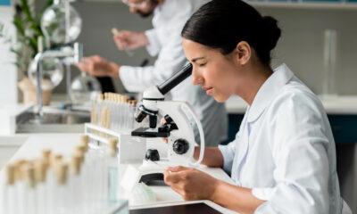 Female scientist in a white lab coat looks through a microscope at a lab bench with glassware in the background.