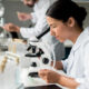 Female scientist in a white lab coat looks through a microscope at a lab bench with glassware in the background.