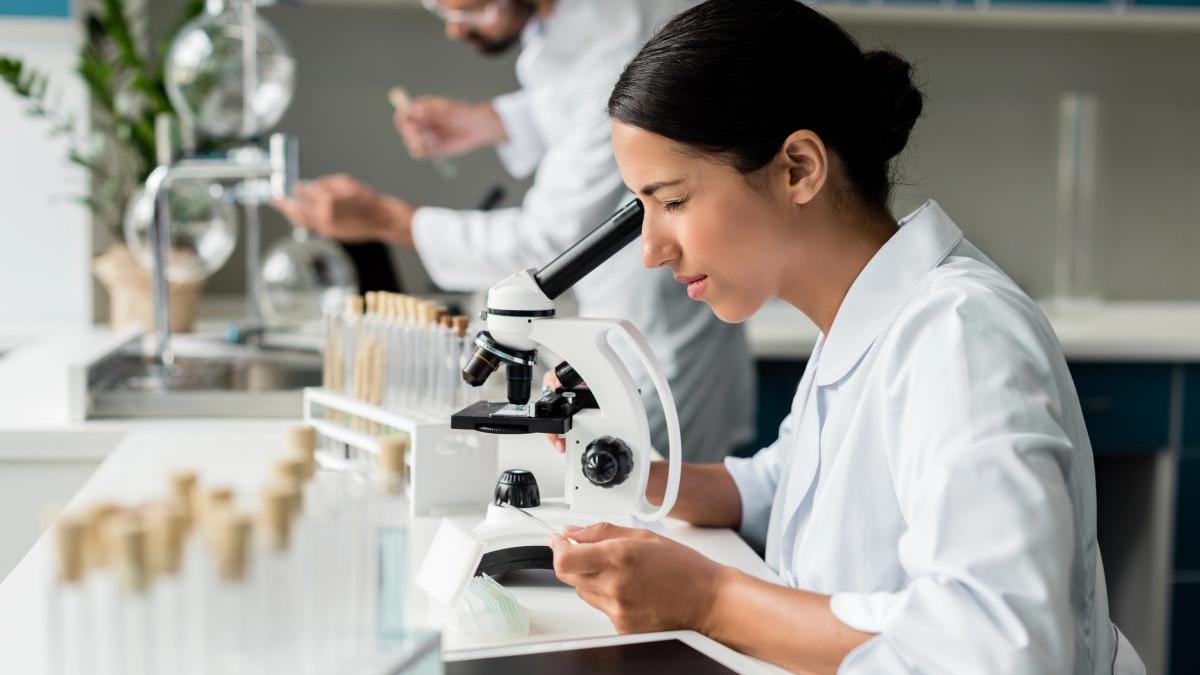 Female scientist in a white lab coat looks through a microscope at a lab bench with glassware in the background.