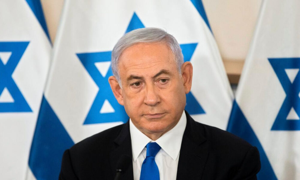 Man in a dark suit and blue tie at a formal press event, with Israeli flags in the background