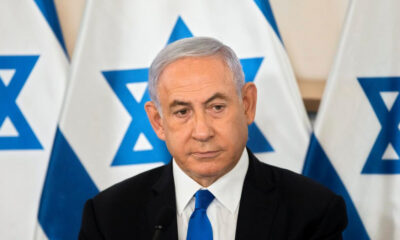 Man in a dark suit and blue tie at a formal press event, with Israeli flags in the background