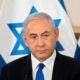 Man in a dark suit and blue tie at a formal press event, with Israeli flags in the background