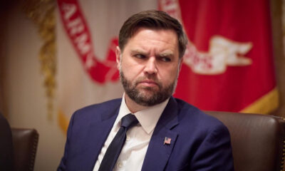 Bearded man in a navy suit and tie with a U.S. flag pin, seated at a formal event