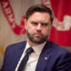 Bearded man in a navy suit and tie with a U.S. flag pin, seated at a formal event