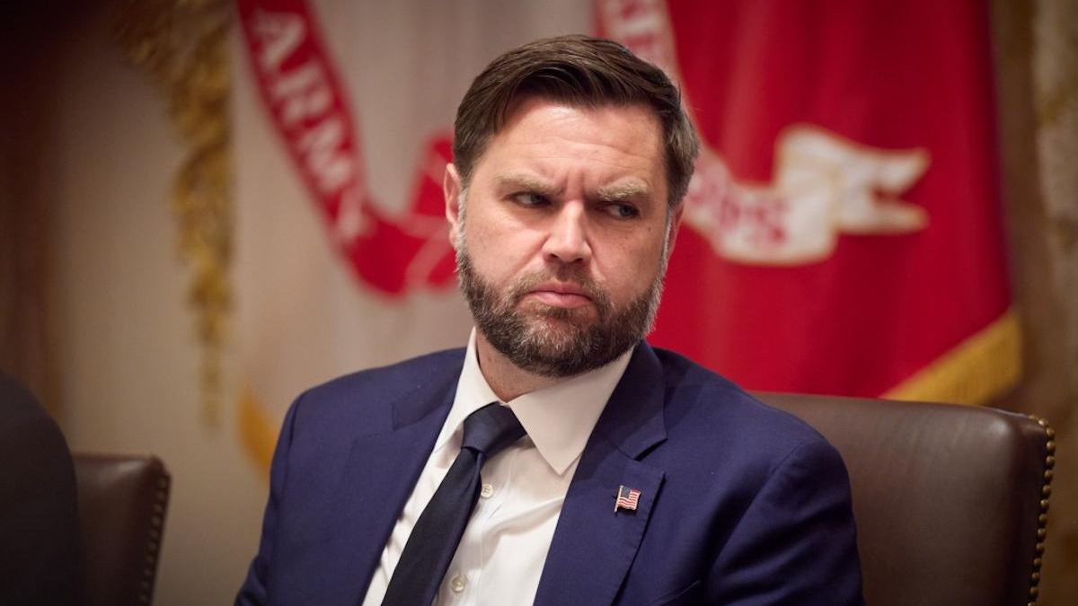 Bearded man in a navy suit and tie with a U.S. flag pin, seated at a formal event