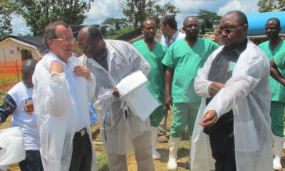 Group of volunteers in protective gowns outside clinic; one man helps another adjust his gown.