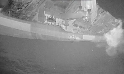 Aerial view of a gray naval ship at sea with a plume of smoke or explosion on the starboard side.