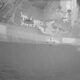 Aerial view of a gray naval ship at sea with a plume of smoke or explosion on the starboard side.