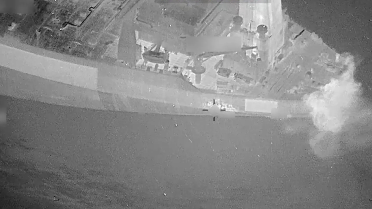 Aerial view of a gray naval ship at sea with a plume of smoke or explosion on the starboard side.