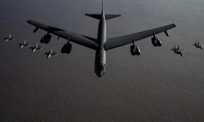 A large military bomber leads a formation of smaller jets flying over a barren, desert-like landscape.