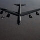 A large military bomber leads a formation of smaller jets flying over a barren, desert-like landscape.
