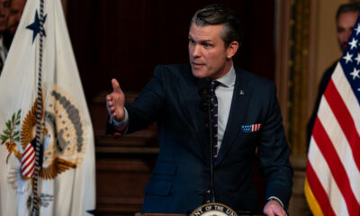 Man in a dark suit speaks at a podium, gesturing with his right hand, with US flags in the background and a government seal emblem on the podium.