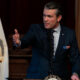 Man in a dark suit speaks at a podium, gesturing with his right hand, with US flags in the background and a government seal emblem on the podium.