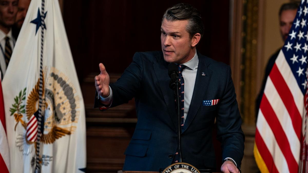 Man in a dark suit speaks at a podium, gesturing with his right hand, with US flags in the background and a government seal emblem on the podium.