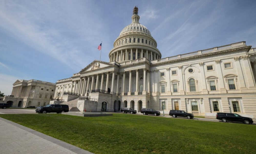 U.S. Capitol building with a large dome, columns, and an American flag, cars parked along the façade on a sunny day.
