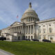 U.S. Capitol building with a large dome, columns, and an American flag, cars parked along the façade on a sunny day.