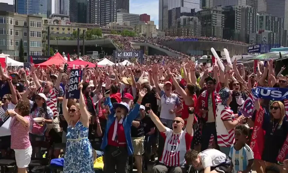 Large crowd of cheering sports fans with arms raised, many wearing red-white-blue clothing and USA banners, in a city waterfront venue.