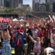 Large crowd of cheering sports fans with arms raised, many wearing red-white-blue clothing and USA banners, in a city waterfront venue.