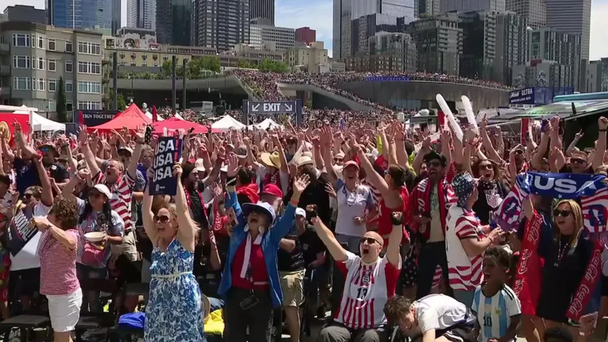 Large crowd of cheering sports fans with arms raised, many wearing red-white-blue clothing and USA banners, in a city waterfront venue.
