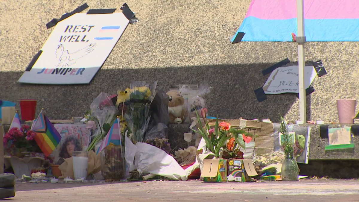 Makeshift memorial on a sidewalk with flowers, candles, signs, and rainbow flags against a concrete wall.