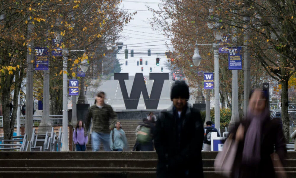 Campus scene featuring a large W sculpture on steps, with fall trees, banners, and pedestrians in the foreground and background on a university campus.