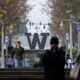 Campus scene featuring a large W sculpture on steps, with fall trees, banners, and pedestrians in the foreground and background on a university campus.