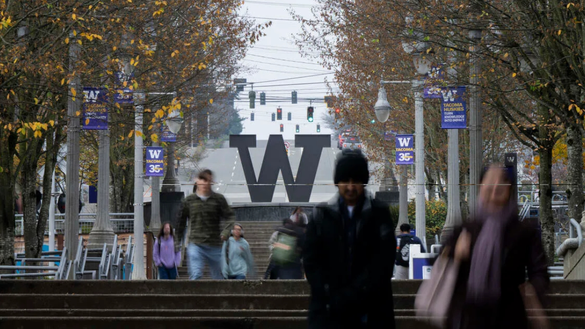 Campus scene featuring a large W sculpture on steps, with fall trees, banners, and pedestrians in the foreground and background on a university campus.