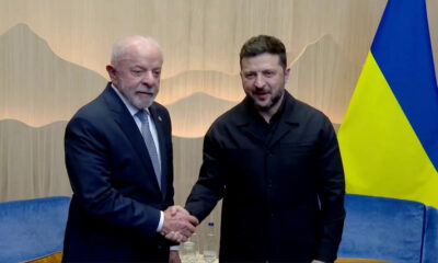 Two men shake hands in a formal meeting, one in a suit and the other in a dark jacket, with a Ukrainian flag behind them.
