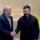 Two men shake hands in a formal meeting, one in a suit and the other in a dark jacket, with a Ukrainian flag behind them.