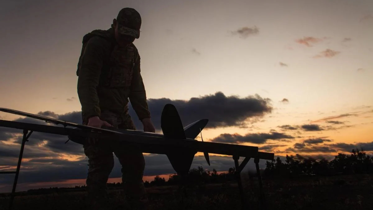Silhouette of a soldier in camouflage adjusting a large unmanned aircraft on a stand at sunset in an open field.
