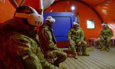 Four soldiers in camouflage with VR headsets seated on chairs inside an orange tent, Ukrainian flag patches on uniforms visible.