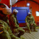 Four soldiers in camouflage with VR headsets seated on chairs inside an orange tent, Ukrainian flag patches on uniforms visible.
