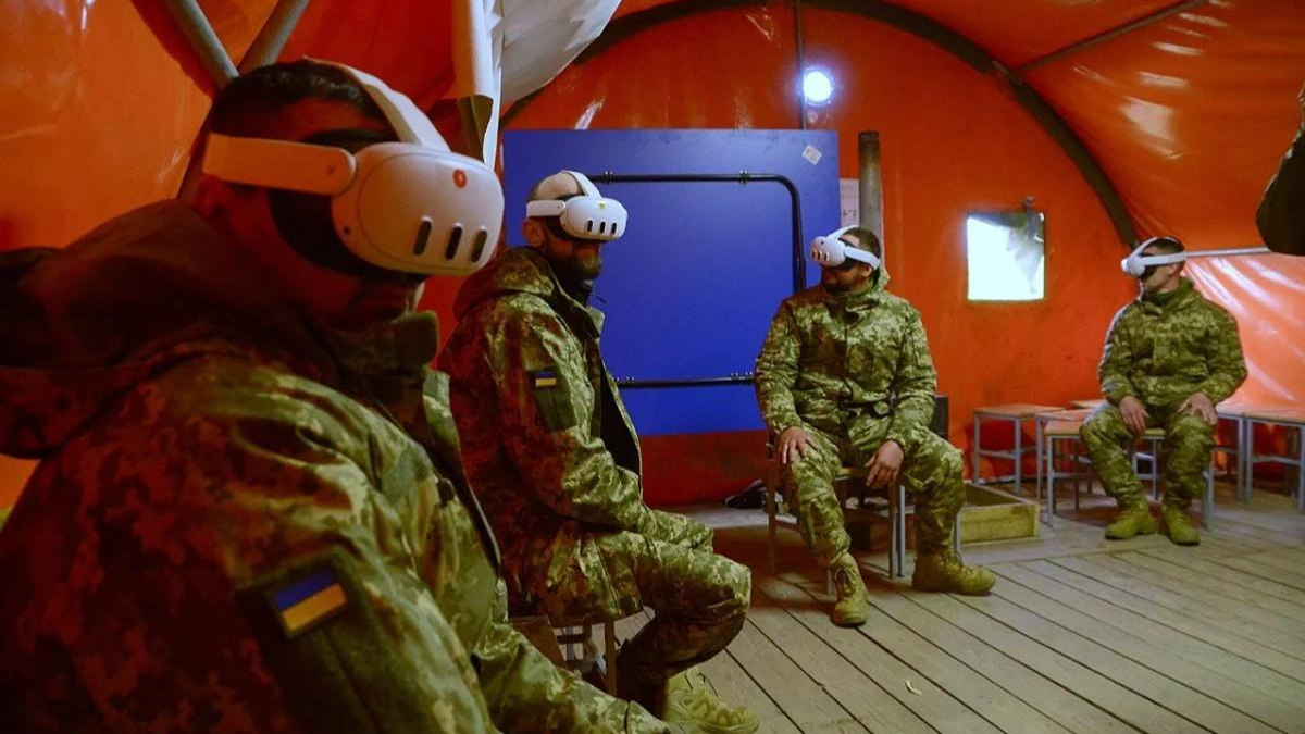 Four soldiers in camouflage with VR headsets seated on chairs inside an orange tent, Ukrainian flag patches on uniforms visible.