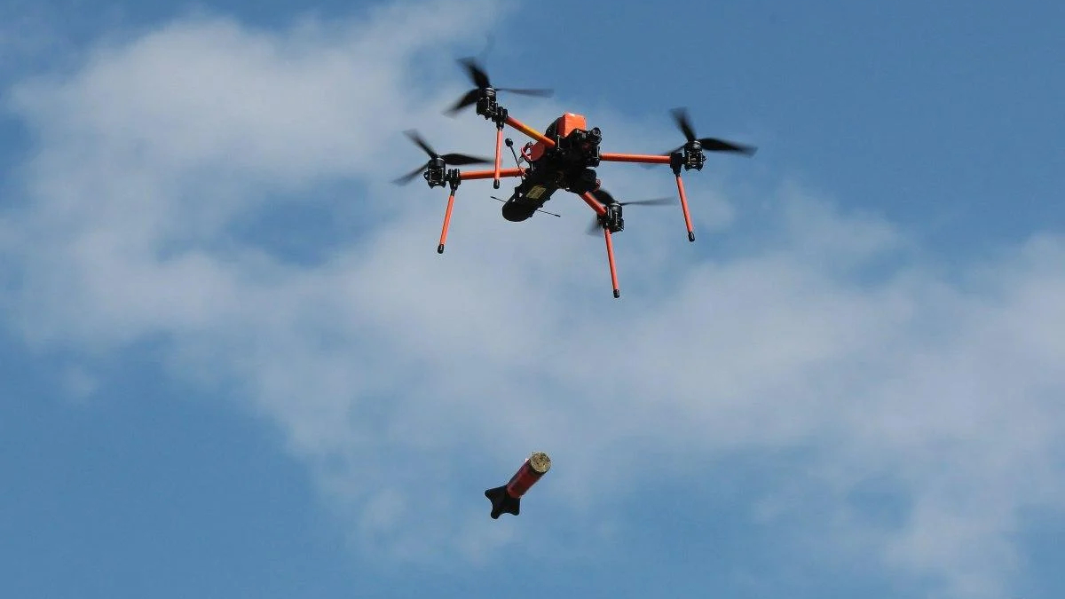 Delivery quadcopter drone releasing a package mid-air against a blue sky with white clouds, orange arms visible in the frame.