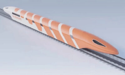 Orange streamlined high-speed train gliding along parallel railroad tracks against a pale sky, viewed from the side with white stripes on its body.