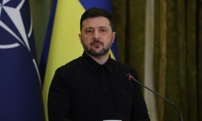 Man in a black shirt speaks at a podium with a microphone, against blue and yellow flags in the background.