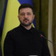 Man in a black shirt speaks at a podium with a microphone, against blue and yellow flags in the background.