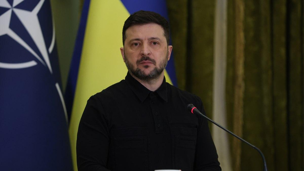 Man in a black shirt speaks at a podium with a microphone, against blue and yellow flags in the background.