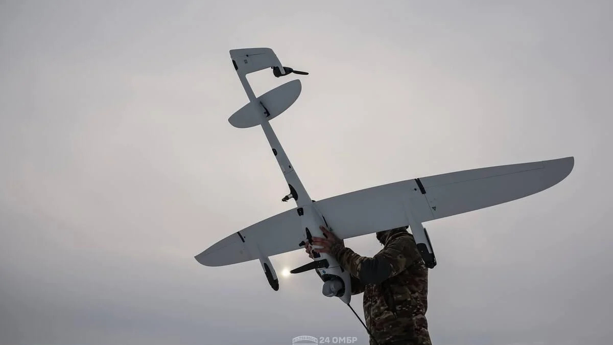 Soldier in camouflage holds a large white fixed-wing unmanned aerial vehicle being prepared for hand launch outdoors.