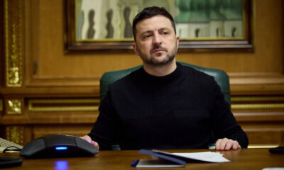 Man in a black sweater sits at a wooden desk in a formal office, with papers and a conference phone in front.