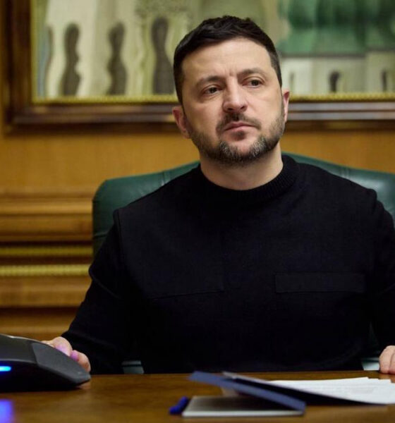 Man in a black sweater sits at a wooden desk in a formal office, with papers and a conference phone in front.