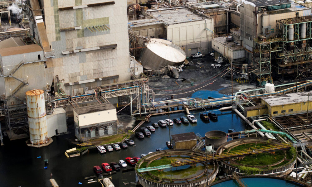 Aerial view of an industrial plant with floodwater surrounding a row of parked cars and large processing equipment nearby to the buildings.