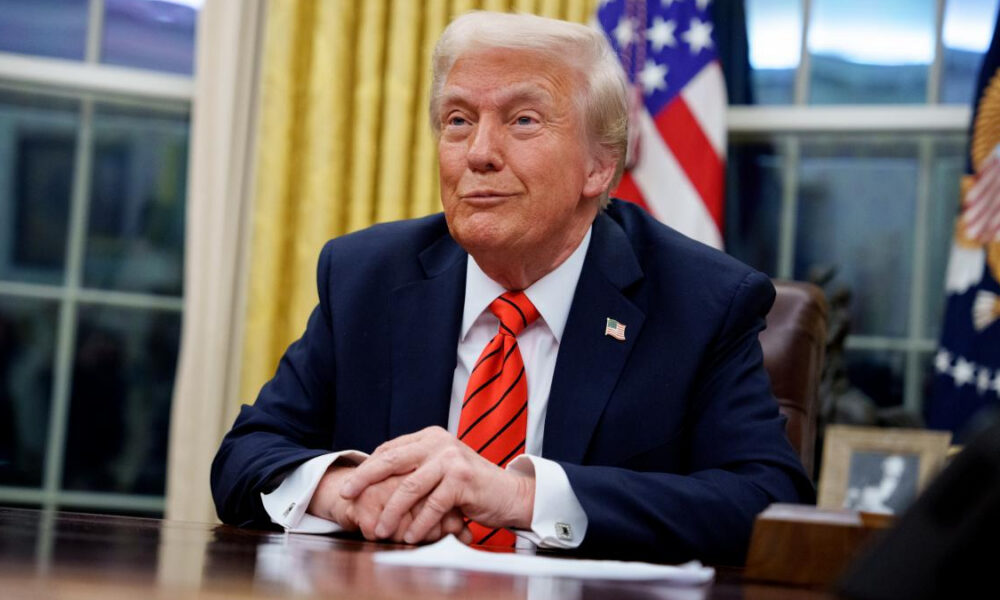 Former U.S. President Donald Trump seated at a desk in the Oval Office, hands folded, with American flag in the background.