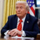 Former U.S. President Donald Trump seated at a desk in the Oval Office, hands folded, with American flag in the background.