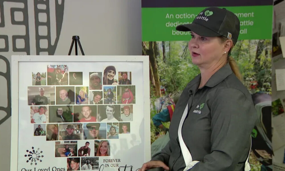 Woman in a dark polo and cap standing beside a photo collage memorial board showing many small portraits; banner reads 'Our Loved Ones' and 'Forever in our hearts'.