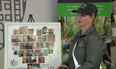 Woman in a dark polo and cap standing beside a photo collage memorial board showing many small portraits; banner reads 'Our Loved Ones' and 'Forever in our hearts'.