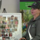 Woman in a dark polo and cap standing beside a photo collage memorial board showing many small portraits; banner reads 'Our Loved Ones' and 'Forever in our hearts'.
