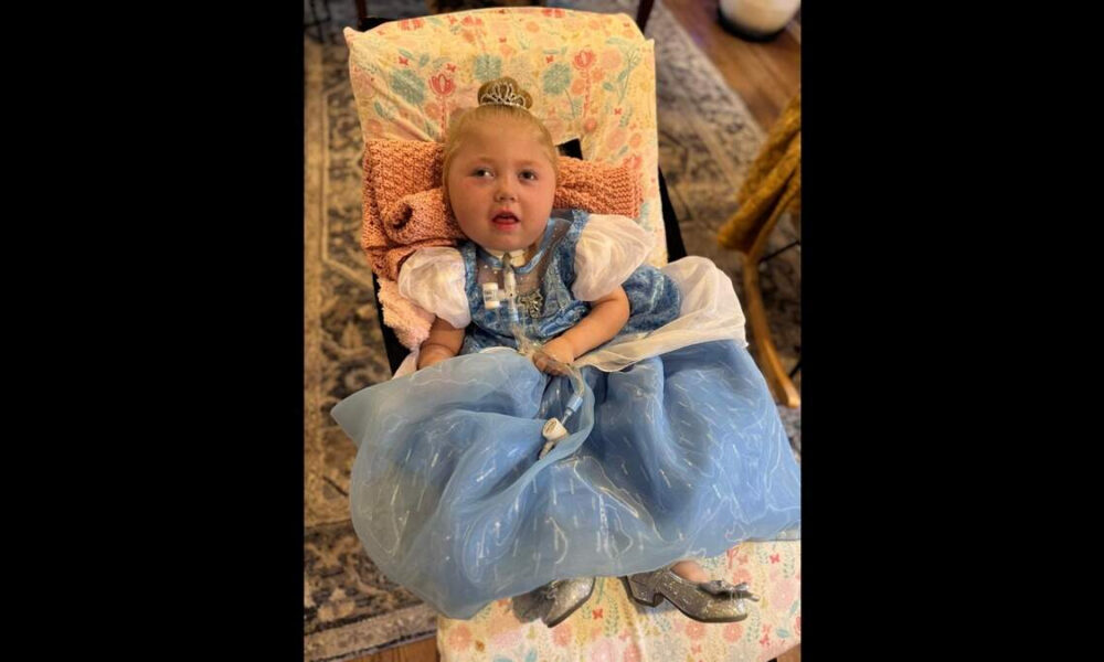 Young girl in a blue princess dress and tiara sitting in a patterned chair, looking toward the camera.