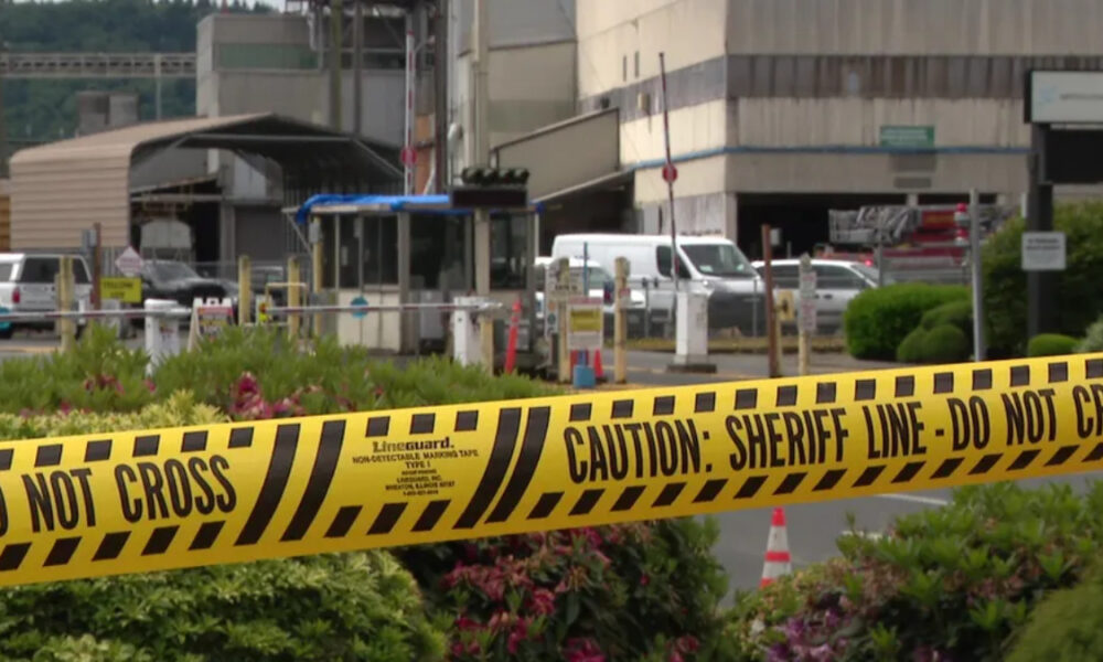 Yellow caution tape reading 'DO NOT CROSS' and 'CAUTION: SHERIFF LINE' in foreground at a restricted area. Industrial buildings and vehicles in background.